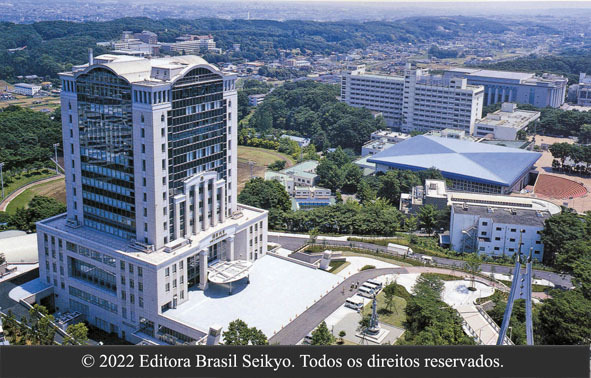 foto de vista aérea do campus da Universidade Soka do Japão, com foco para o prédio mais alto de cor cinza claro, envolto por árvores, ruas e demais construções mais baixas