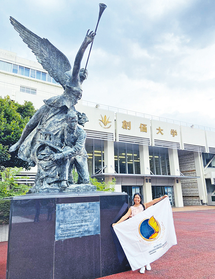 Segura a bandeira da Divisão dos Universitários (DUni) à frente de um dos prédios da Universidade Soka nas terras do Mestre.