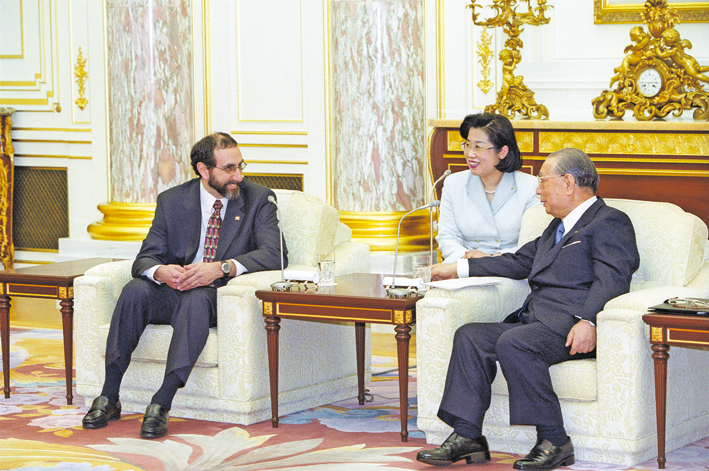 foto de três pessoas sentadas, duas delas sorridentes, viradas uma em direção à outra, à frente e sentados em poltronas brancas: à esquerda, Lou Marinoff, de terno escuro, e Daisaku Ikeda à direita