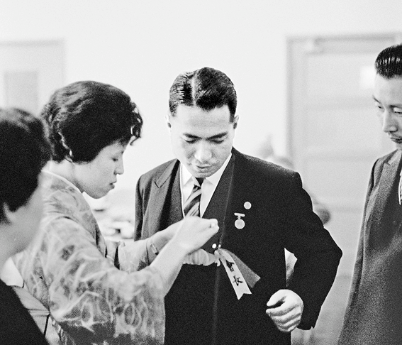 sua esposa, Kaneko, coloca o oribon (acessório em formato de flor feito com fitas) na lapela do fraque dele antes da cerimônia (3 maio 1960)